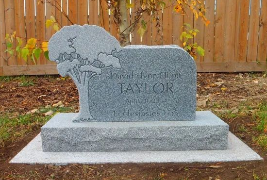 Gray memorial headstone with tree engraving in a yard, set on a raised base before a wooden fence.