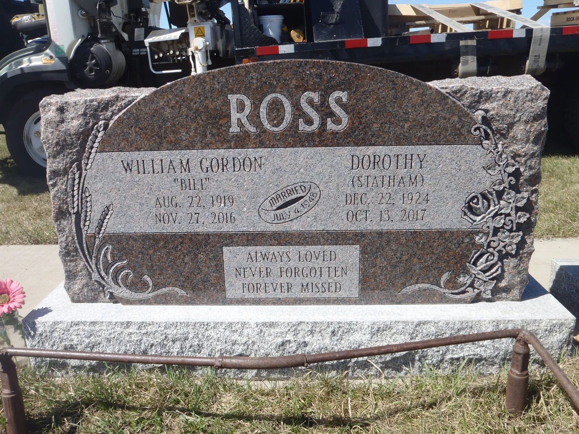 Granite gravestone with “ROSS” engraved, set outdoors on a grassy plot with flowers nearby.