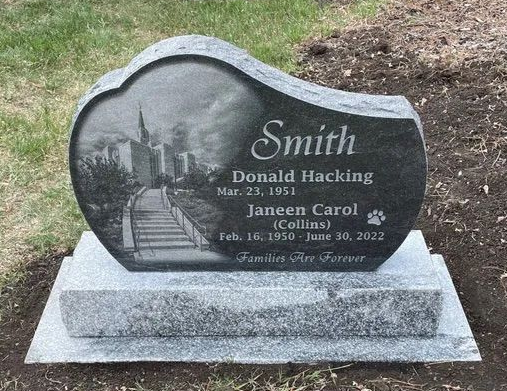Gray heart-shaped gravestone with engraved names and a city skyline, set on a concrete base in grass.