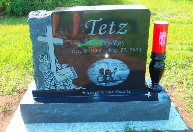 Black granite gravestone with a cross, bicycle engraving, and red candle in a sunny cemetery