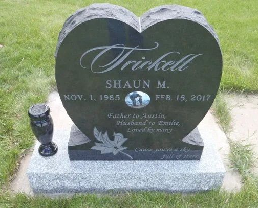 Heart-shaped black granite gravestone with engraved text on a gray base in a grassy cemetery.