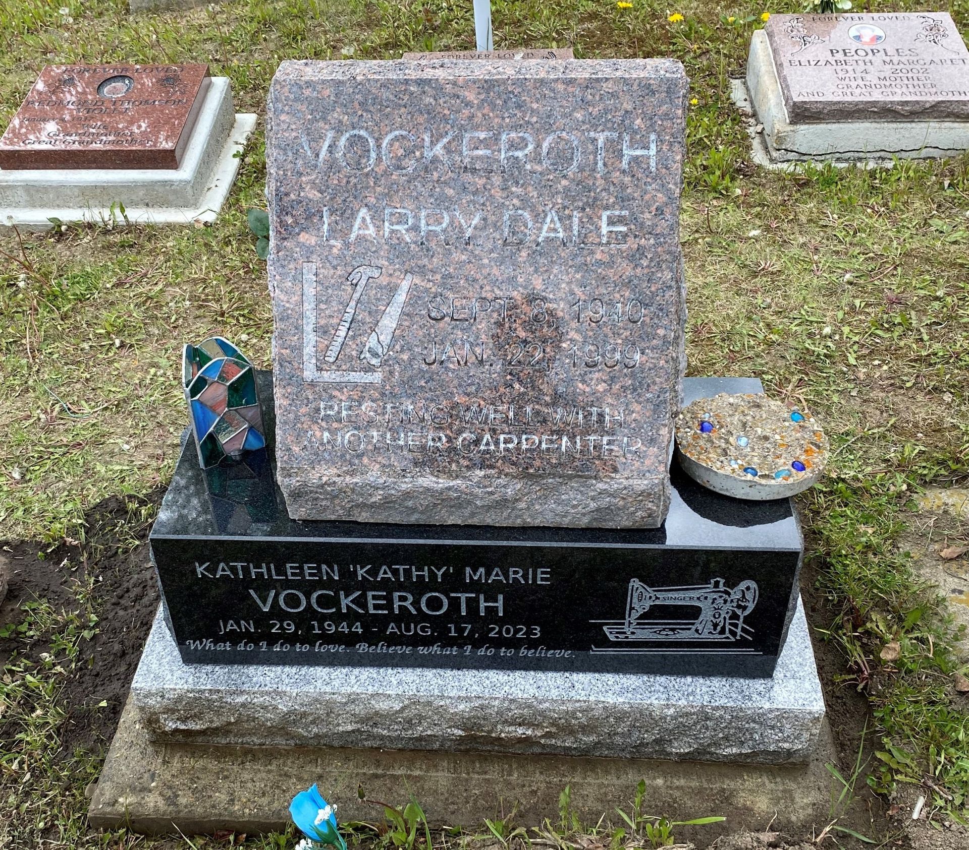 Gravestone with engraved name on gray stone, flowers and a small memorial item on the base in a grassy cemetery.
