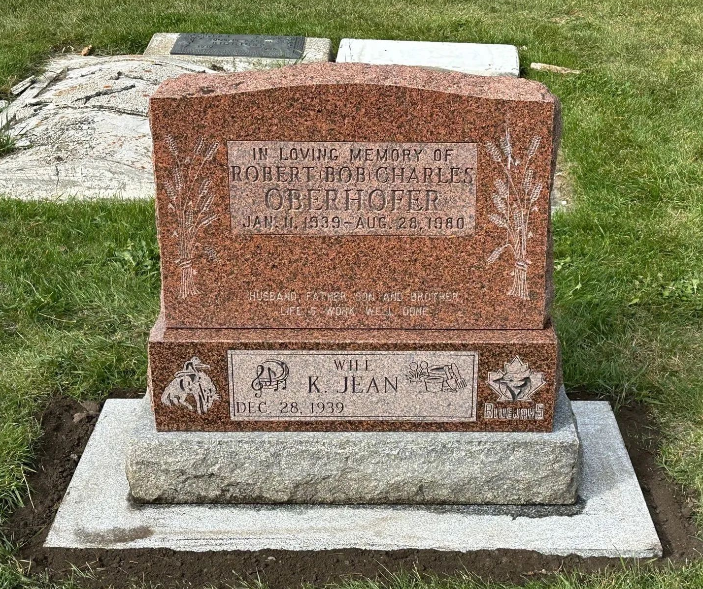Granite gravestone with engraved plaque on a concrete base in a grassy cemetery.