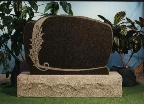 Large dark stone monument with a floral relief, set on a rough beige base outdoors among plants