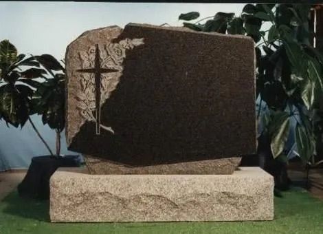 Stone monument with carved cross on a grassy base, framed by plants outdoors