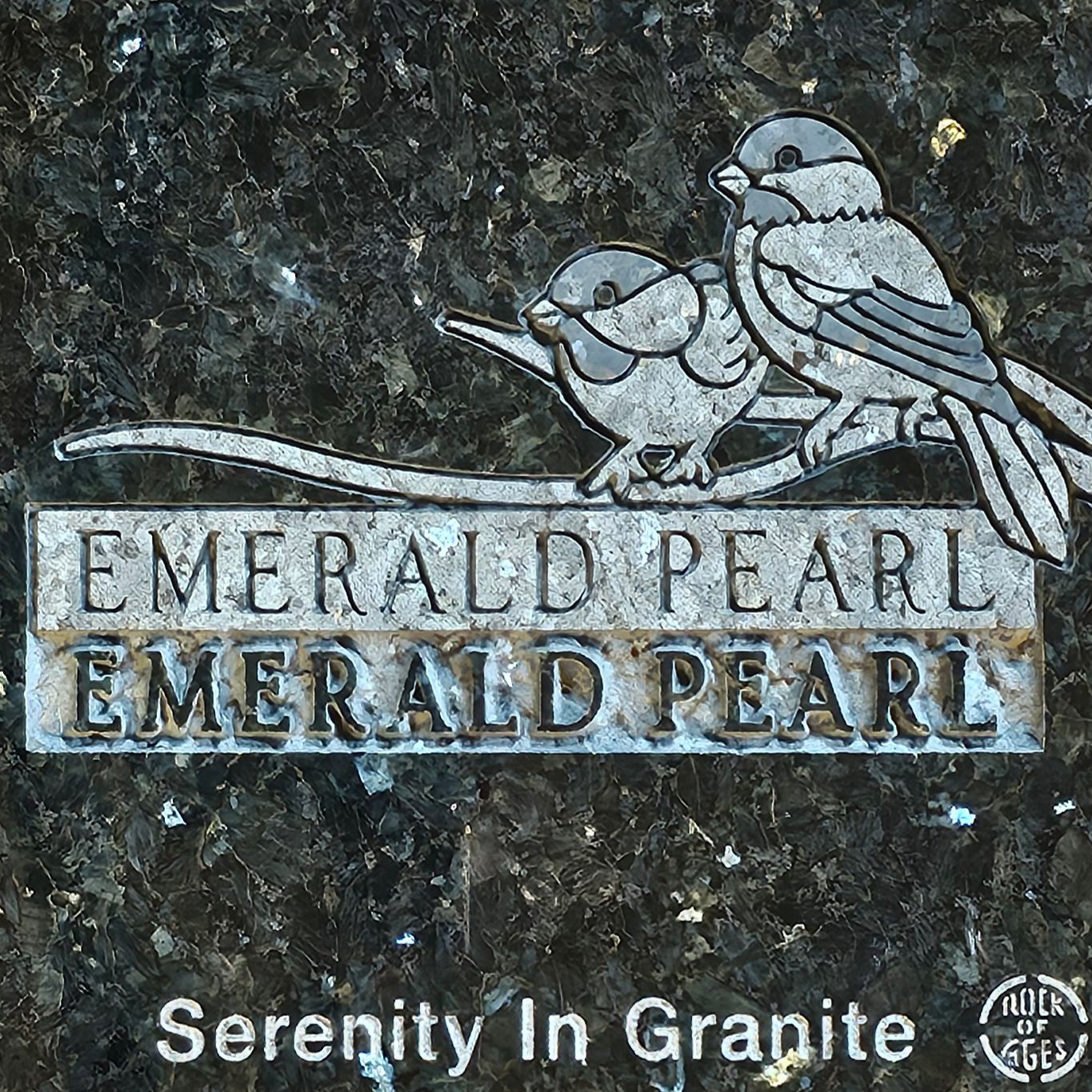 Stone sign reading “EMERALD PEARL” with two birds perched on top and “Serenity in Granite” below.