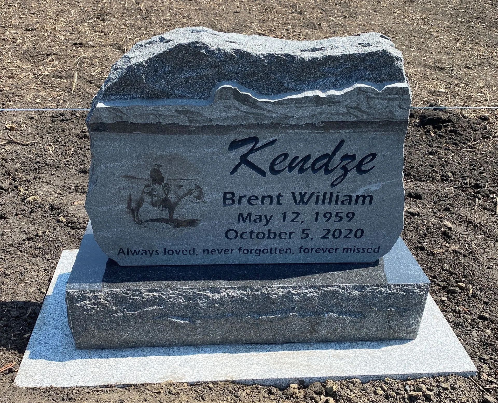 Gray gravestone in a cemetery, engraved with “Kendze” and “Brent William,” on a white base.