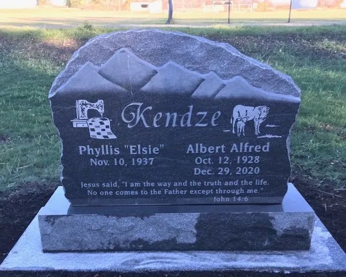 Black granite grave marker with “Kendze” and carved sheep in a grassy cemetery.