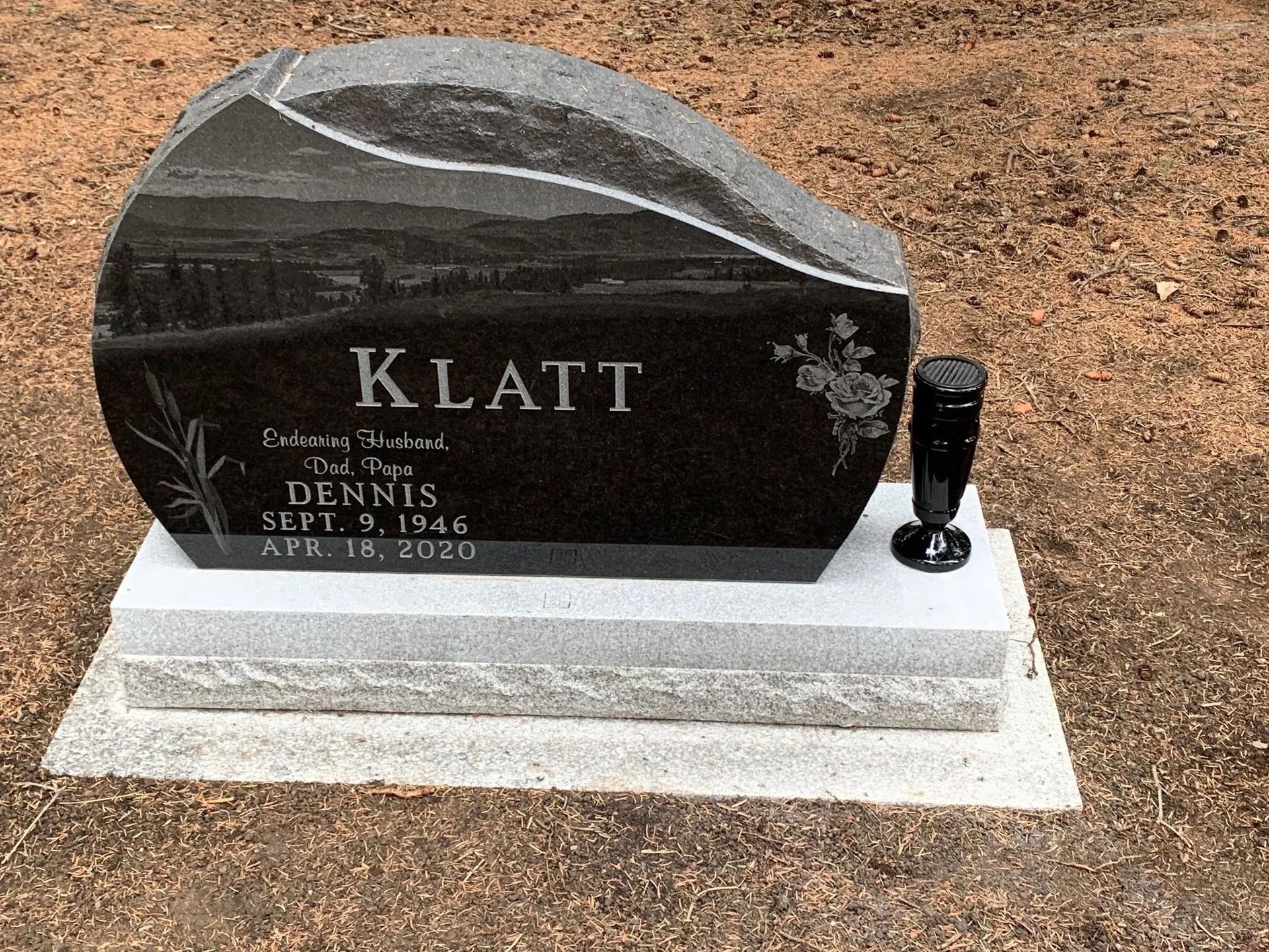 Black granite gravestone with the name “KLATT” on a white base in a dirt cemetery.