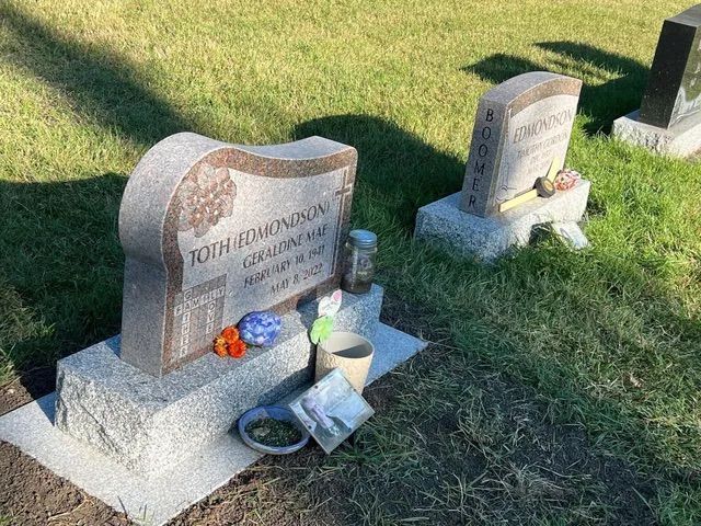 Two granite gravestones with flowers and memorial items on a grassy cemetery plot.