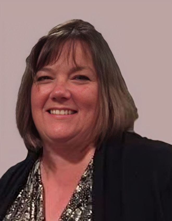 Smiling woman in a dark blazer and patterned top against a plain beige background