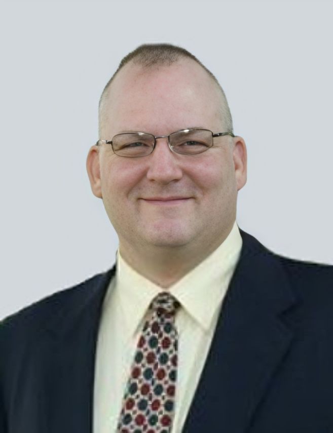 Portrait of a man in glasses and a suit with a patterned tie, smiling against a light background