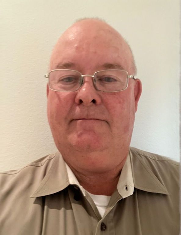 Man wearing glasses and a beige shirt, facing the camera against a plain wall.