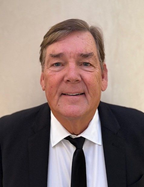 Man in a black suit and tie, smiling against a plain beige background.