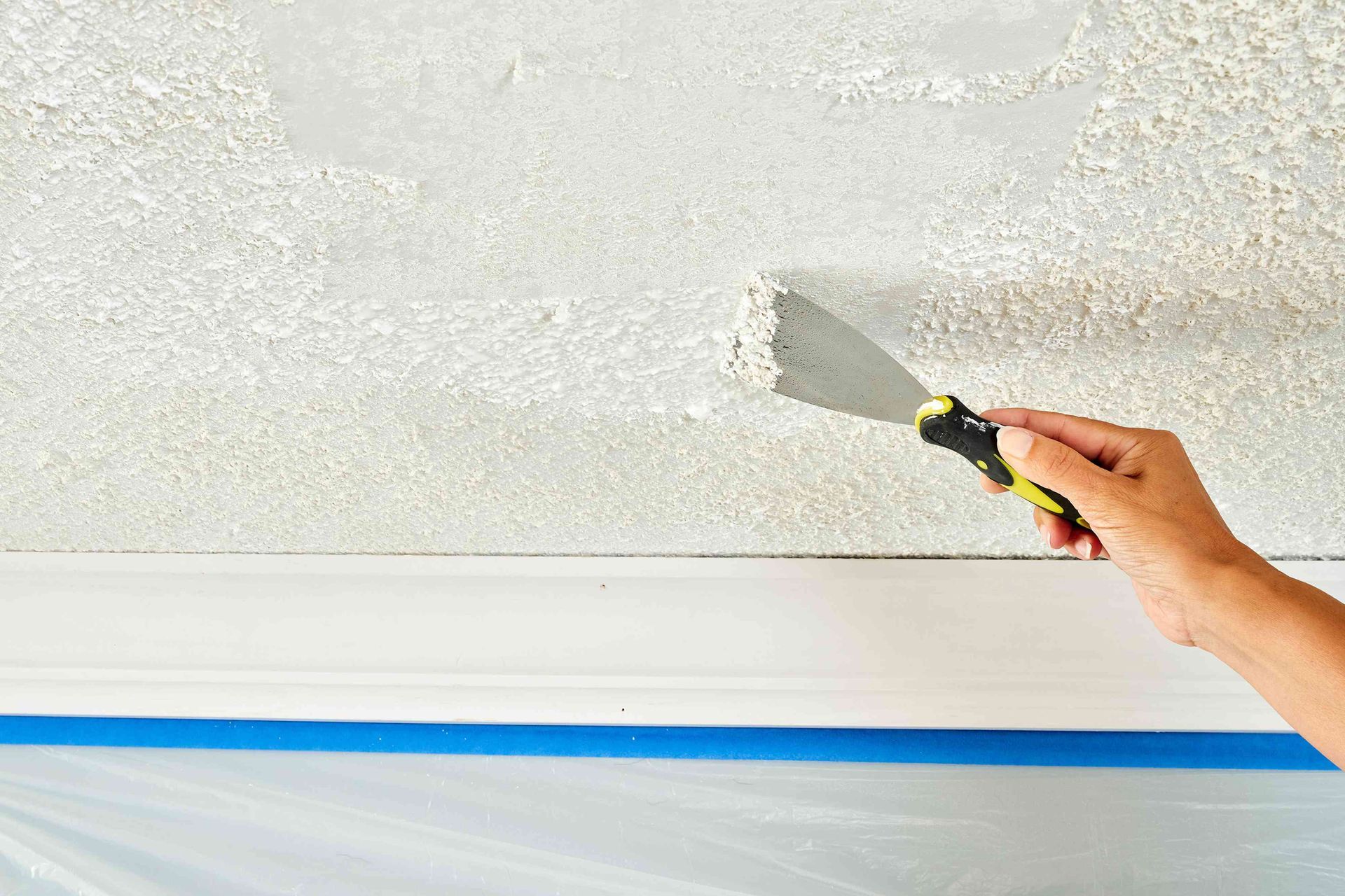 Hand using a putty knife to scrape textured white ceiling near a taped wall edge