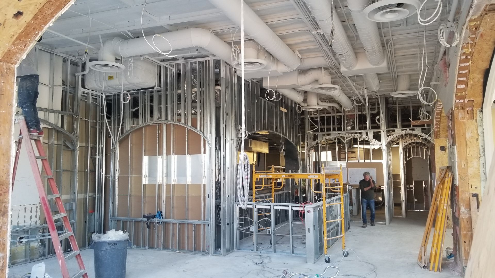 Interior of a building under renovation, with metal framing, scaffolding, exposed ducts, and a worker in the background