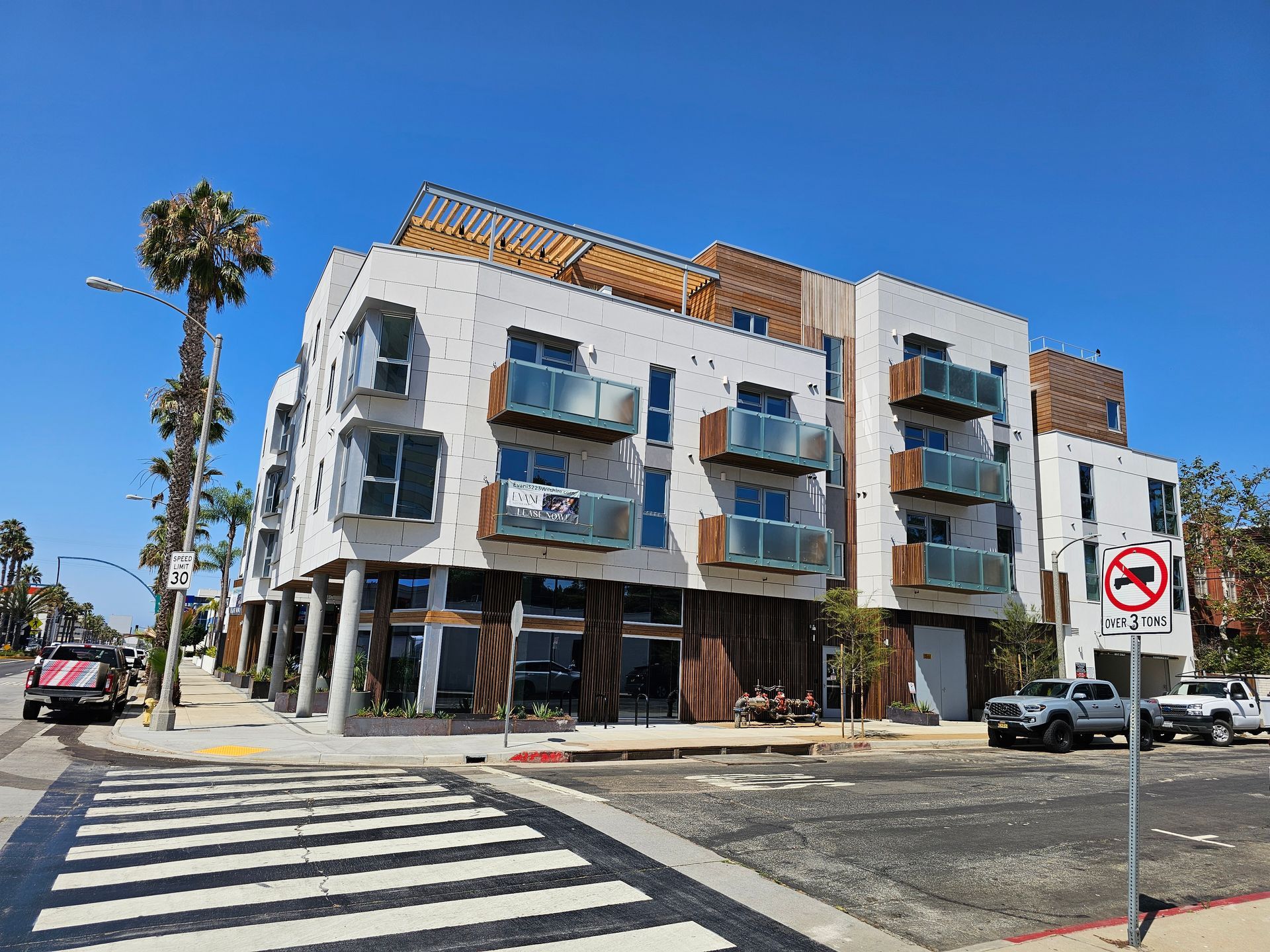 Modern apartment building with balconies at a street corner, palm trees, and a crosswalk under a clear sky