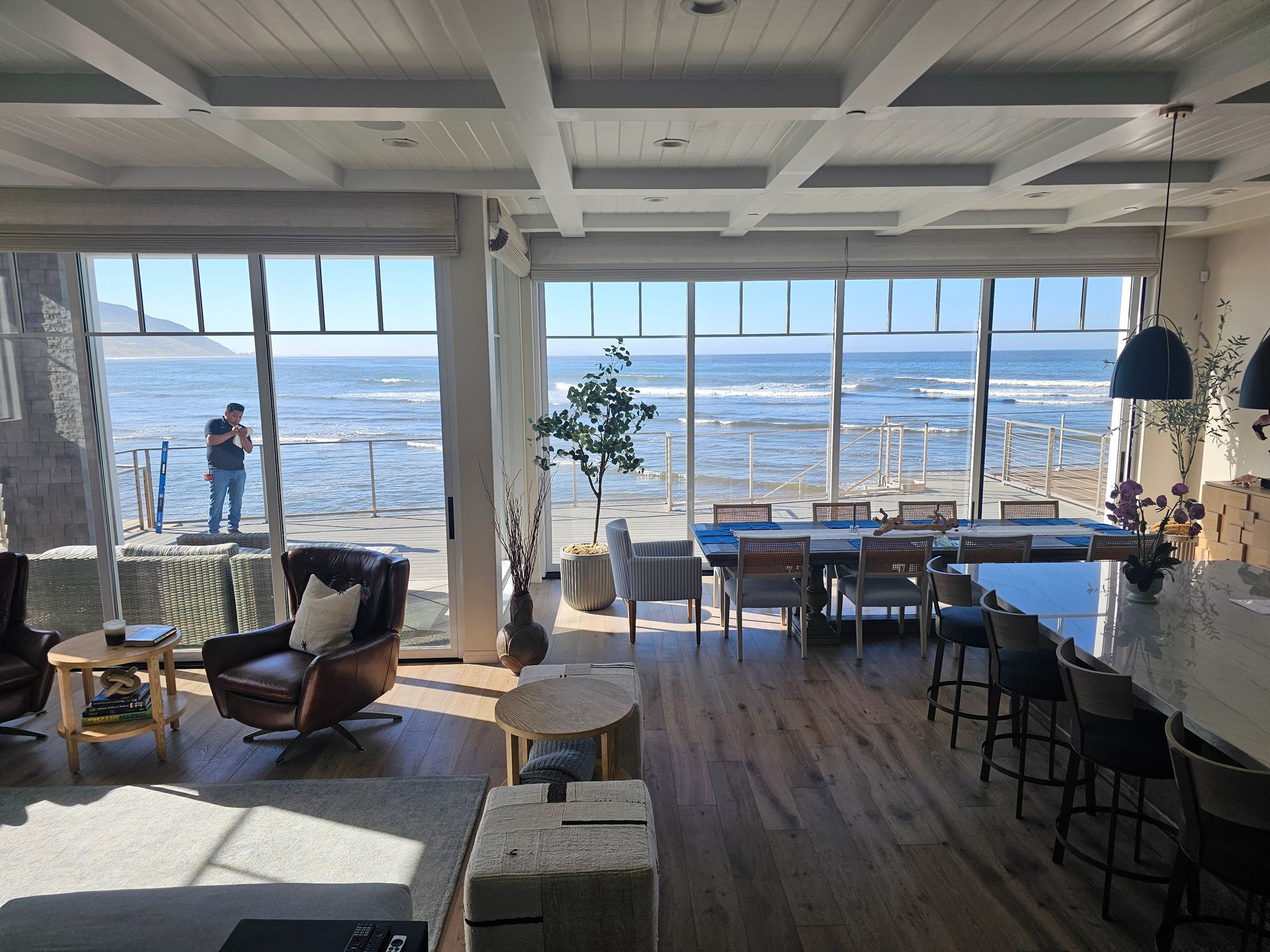Bright seaside café interior with large windows, ocean view, and a few people seated near the shore