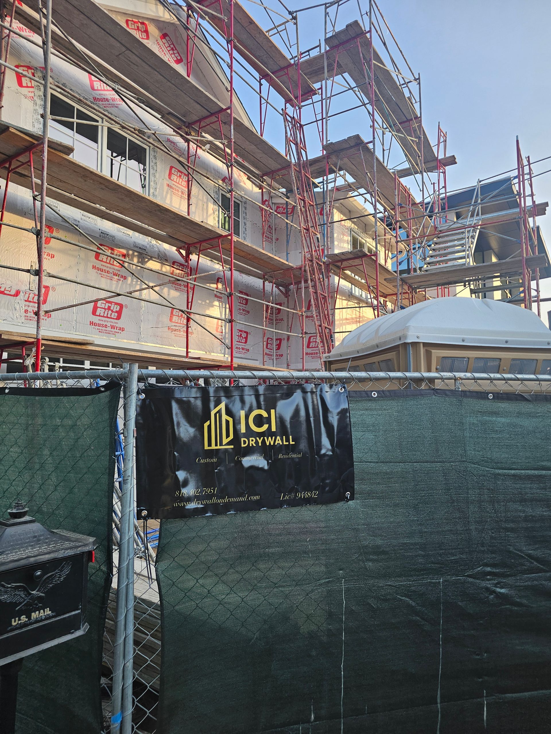 Scaffolding around a building under construction, with green fencing and a black banner in front.