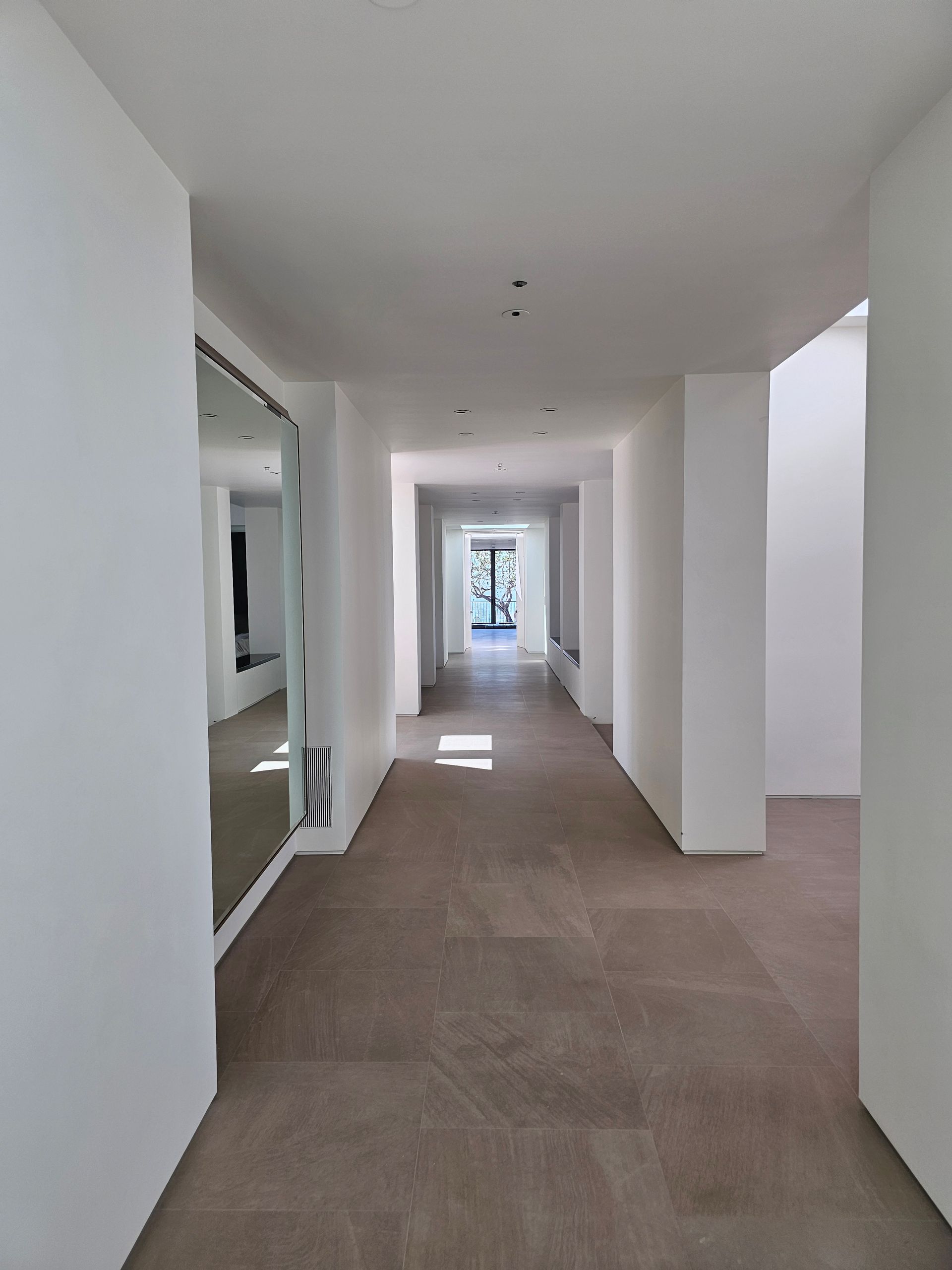Bright empty hallway with white walls, brown carpet, and a window at the far end