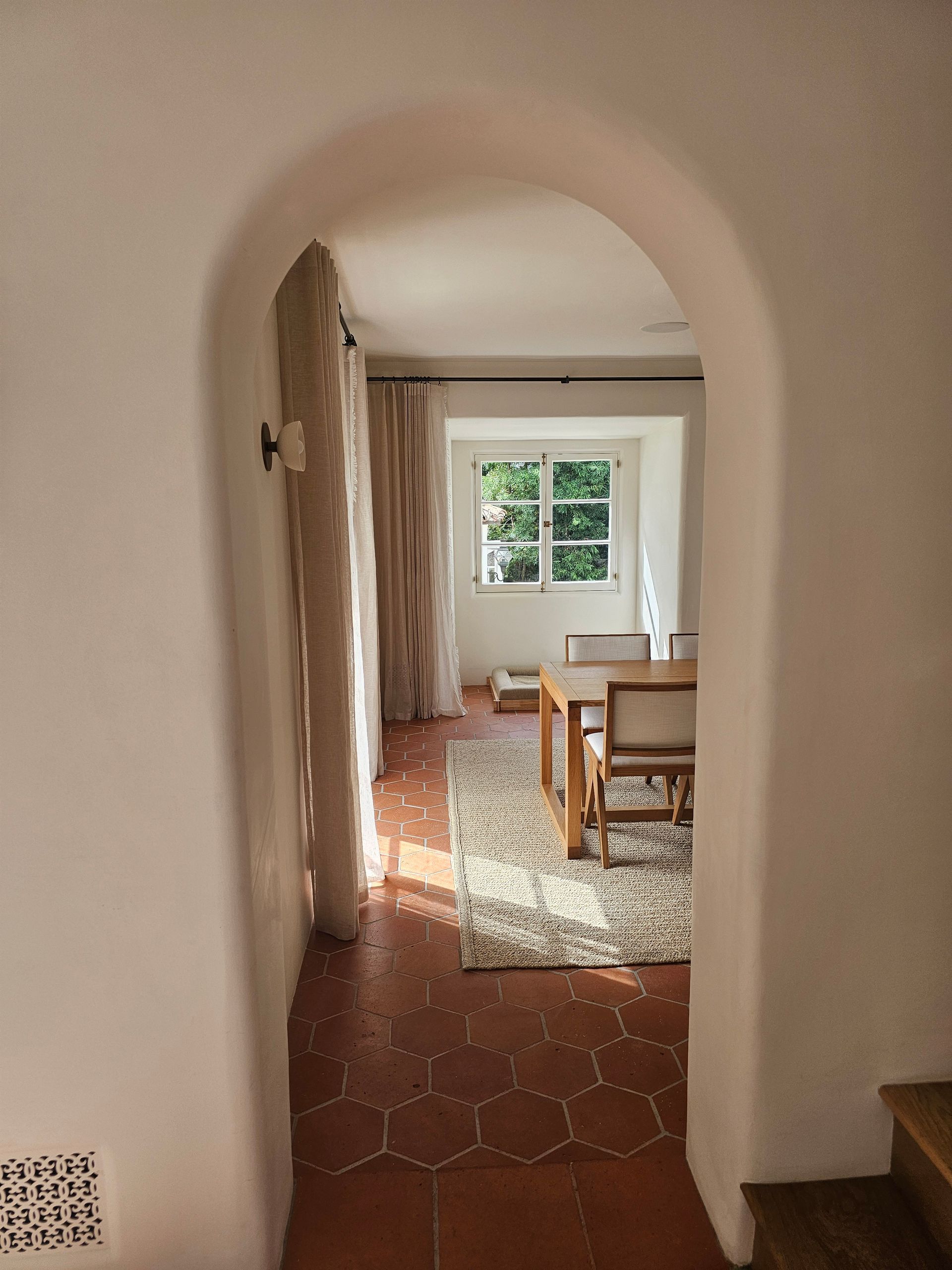 Arched hallway leads to a sunlit room with a table, chair, rug, and window.