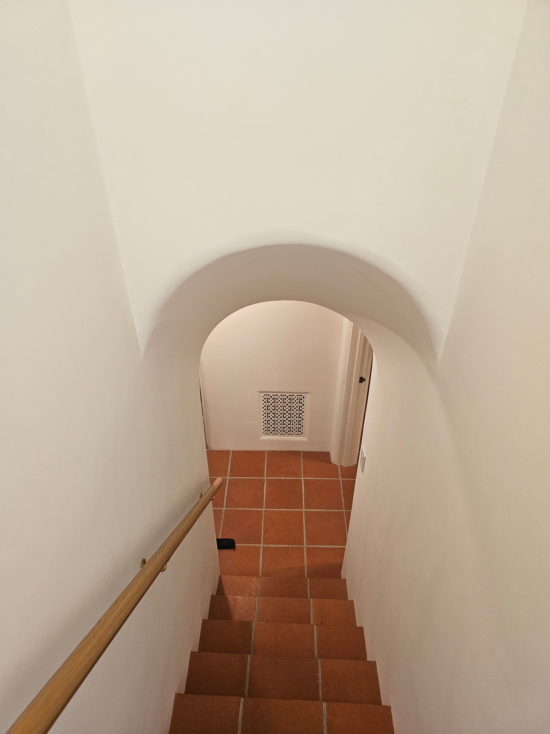 Narrow white stairwell with terracotta tile steps and a wooden handrail, leading down to a small arched doorway