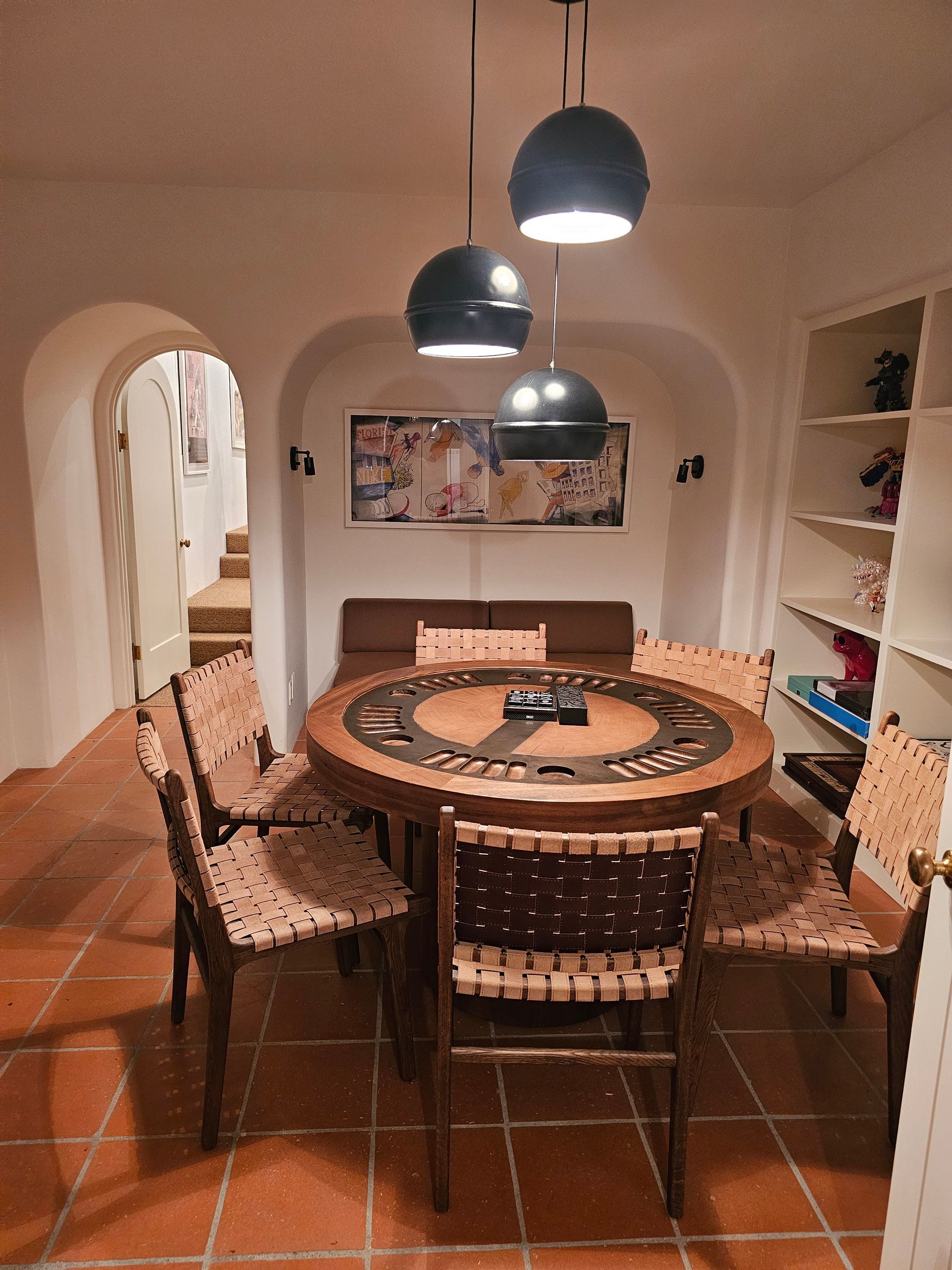 Dining room with round wooden table, patterned chairs, hanging black lamps, and terracotta tile floor.