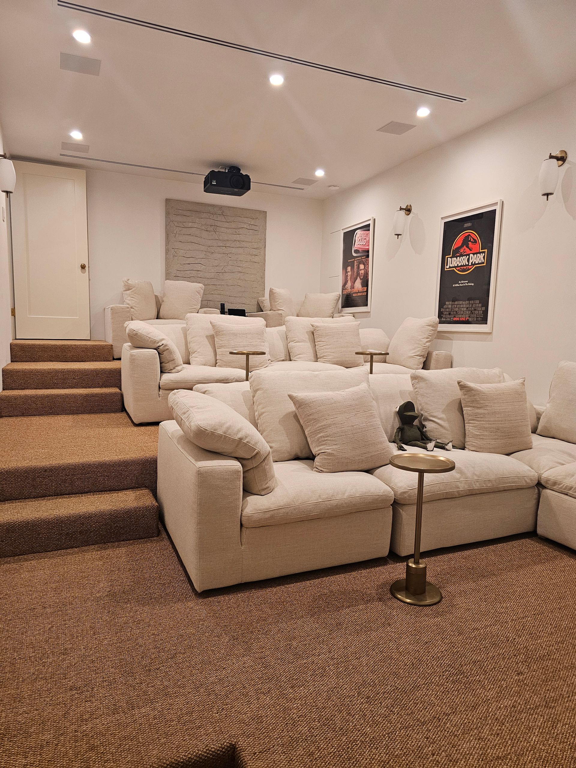 Cozy home theater with white sectional sofa, plush chairs, wall art, and carpeted steps leading up to a projection screen.