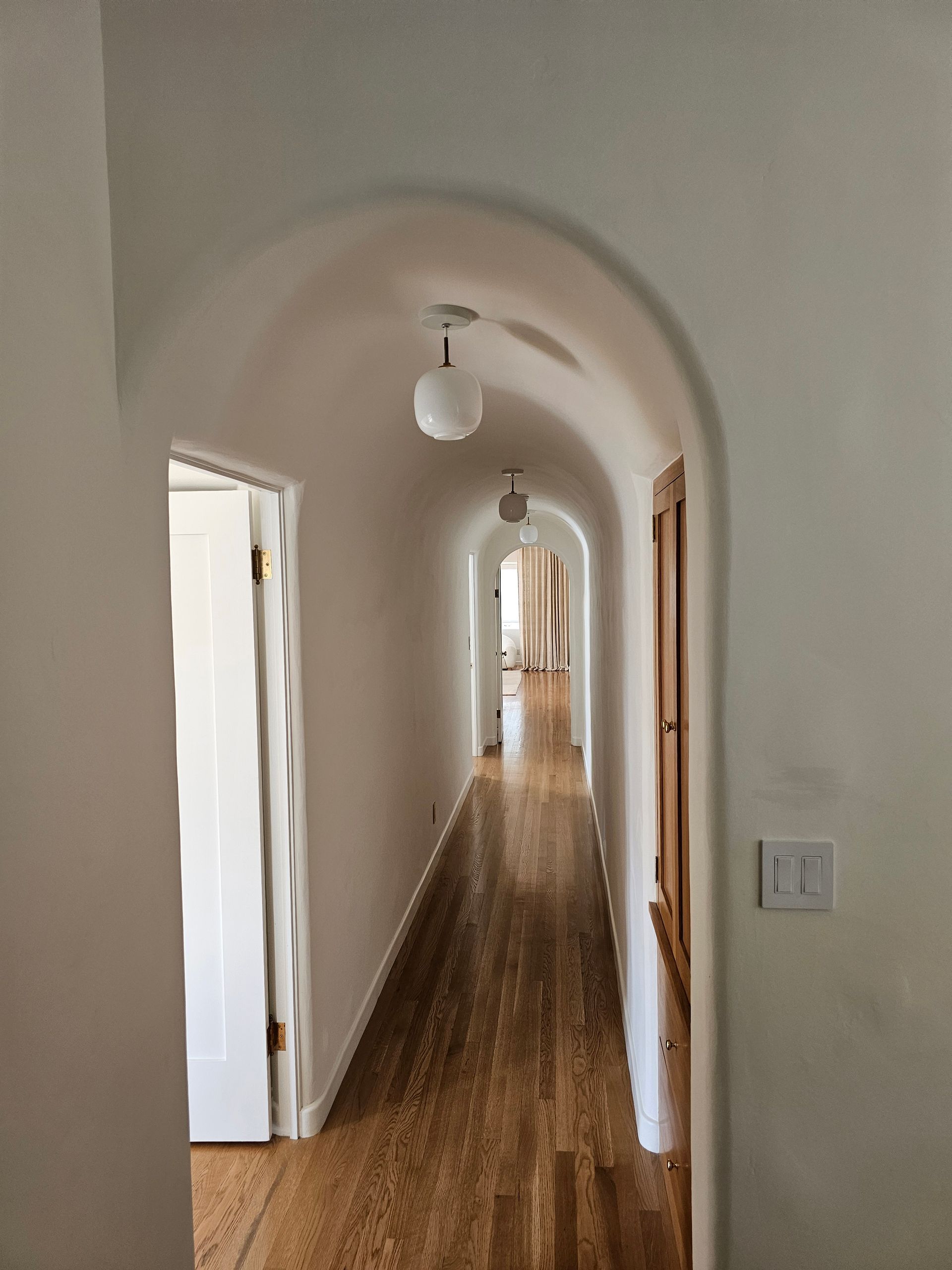 Long arched hallway with wood floor, white walls, hanging lights, and doors along both sides