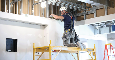 Worker on yellow scaffolding installing ceiling materials in a bright construction site.