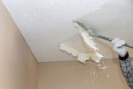 Person scraping damaged popcorn ceiling with a putty knife in a beige room