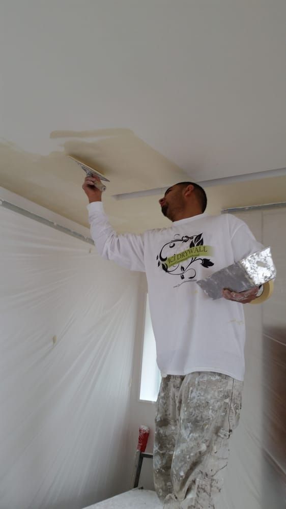 Person painting a ceiling with a roller in a room covered in plastic sheeting
