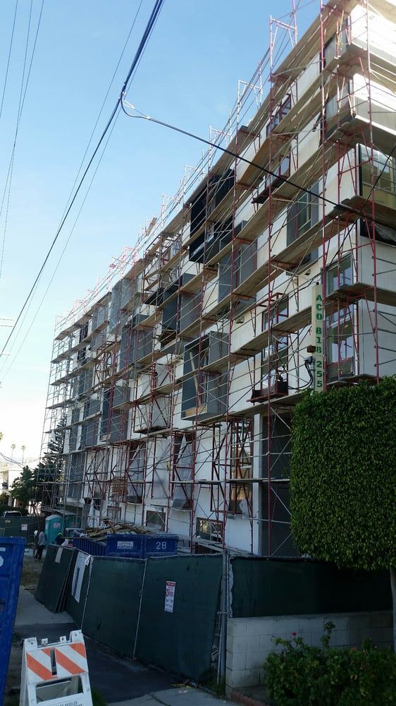 Scaffolding covers a multi-story apartment building under renovation beside a sidewalk and fence.