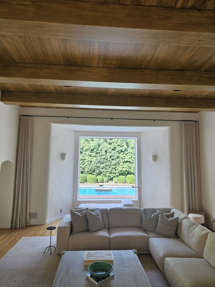 Bright living room with beige sectional, wooden ceiling, and a window overlooking a pool and greenery