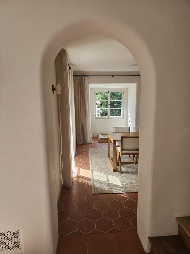 Arched hallway opening into a sunlit room with a table, chair, window, and curtains.