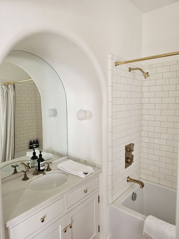 Small white bathroom with vanity sink, arched mirror, and tiled shower with tub and gold fixtures.