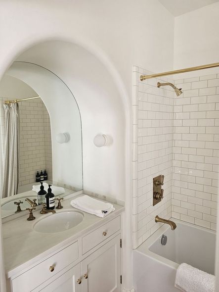Small white bathroom with vanity sink, arched mirror, and tiled shower with tub and gold fixtures.