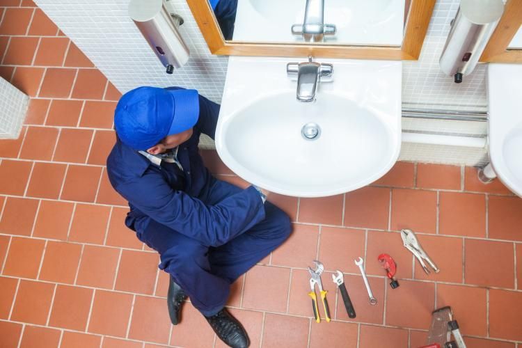 A worker in a blue uniform sits on a tiled floor, repairing a bathroom sink with tools laid out nearby.