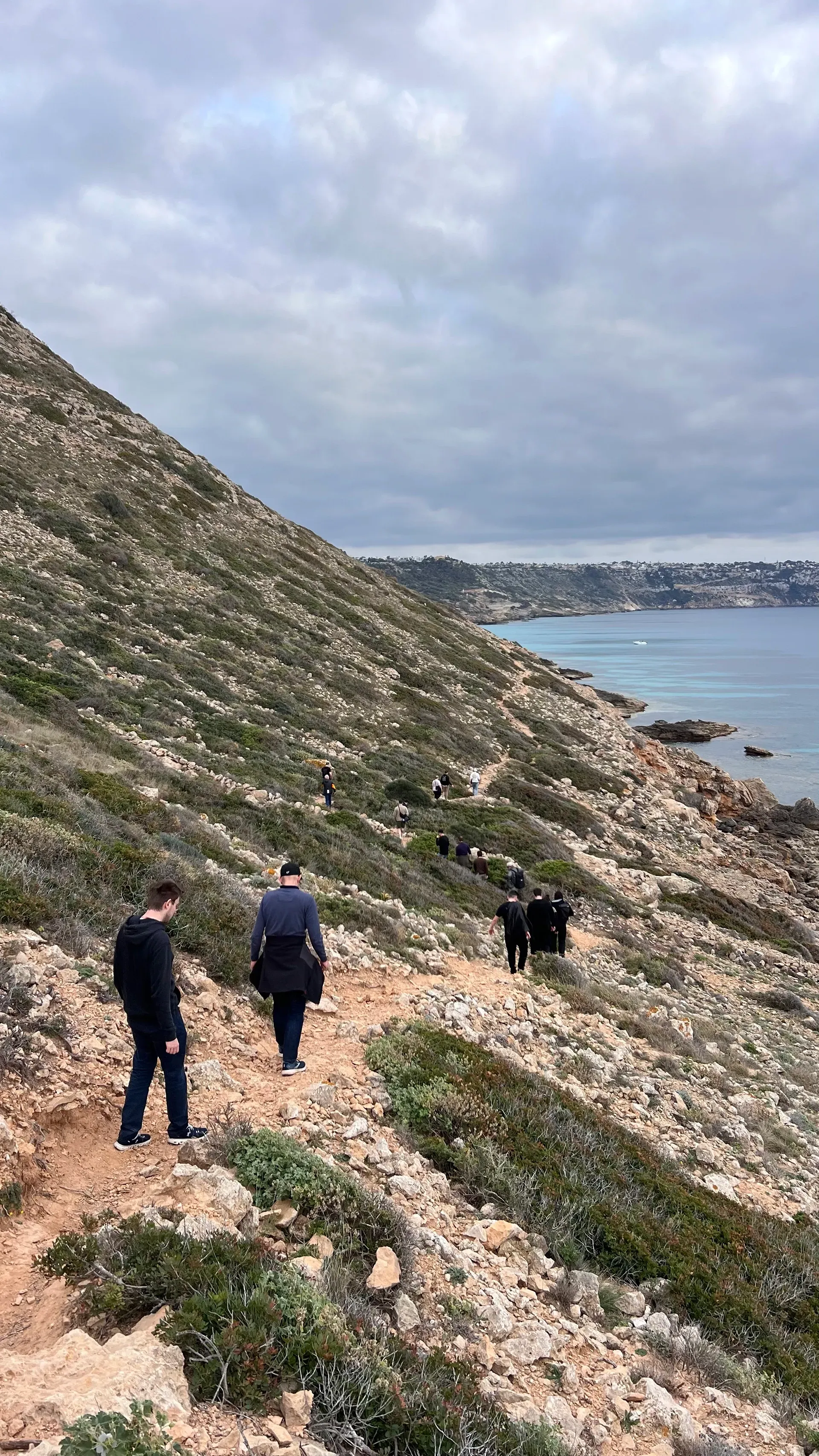 Drei Personen wandern auf einem felsigen Küstenpfad entlang, links erhebt sich ein steiler Hügel, und in der Ferne ist das Meer zu sehen.