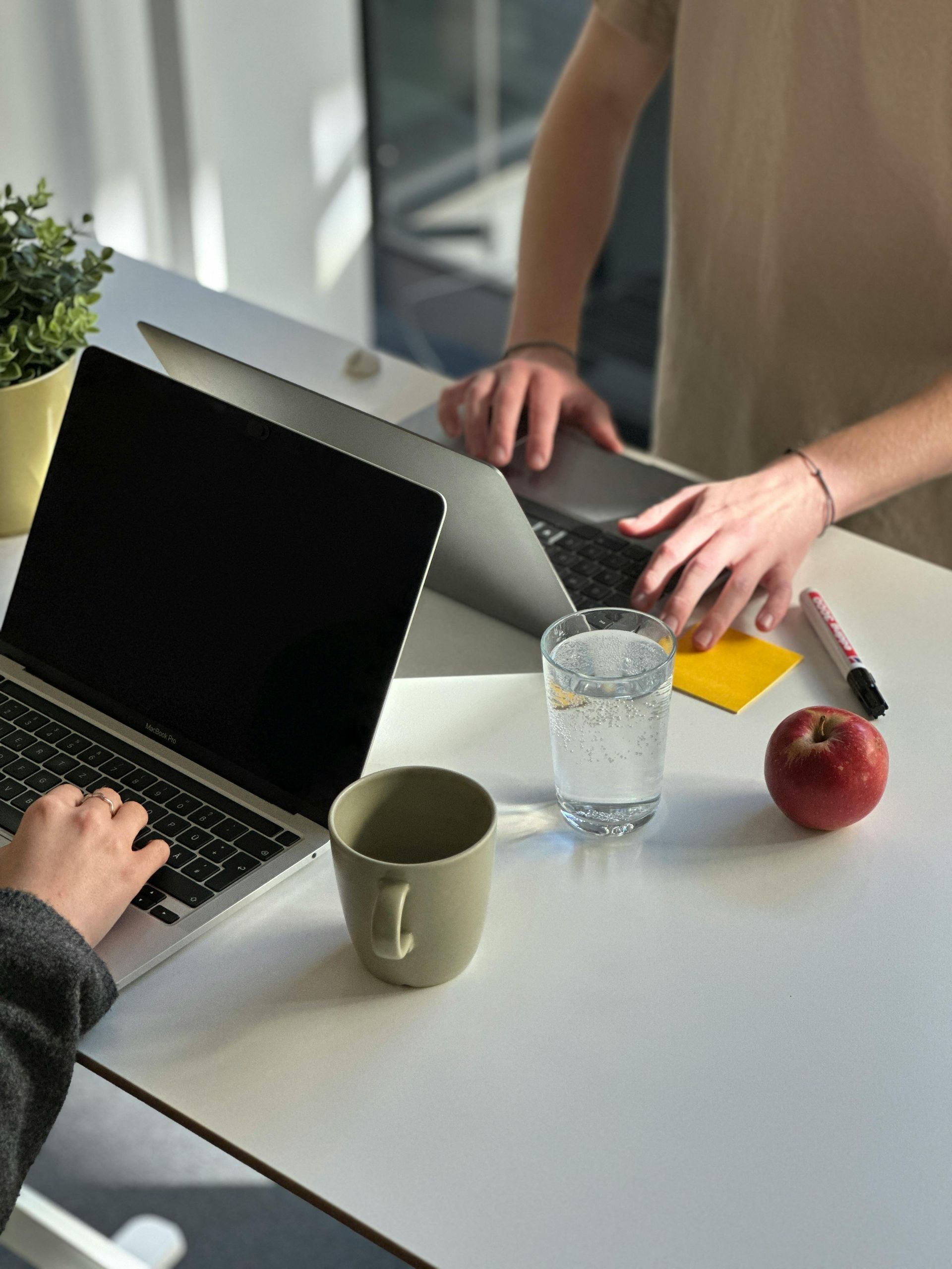 Zwei Personen arbeiten an getrennten Laptops an einem weißen Schreibtisch. Neben ihnen stehen eine Tasse, ein Glas Wasser, ein gelber Zettel und ein roter Apfel.