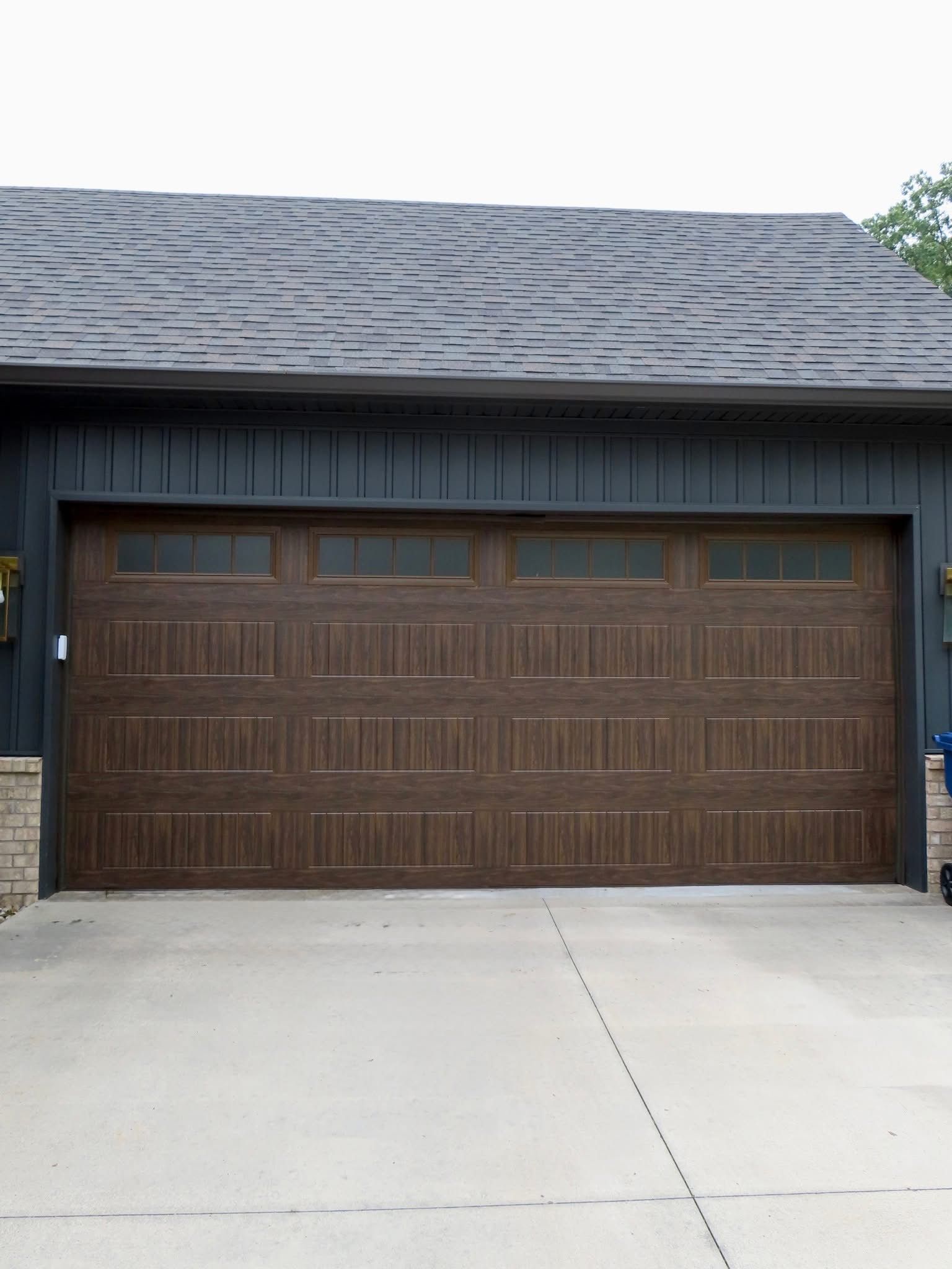 Brown garage door with windows, in front of a blue-gray house with a dark roof and a concrete driveway.