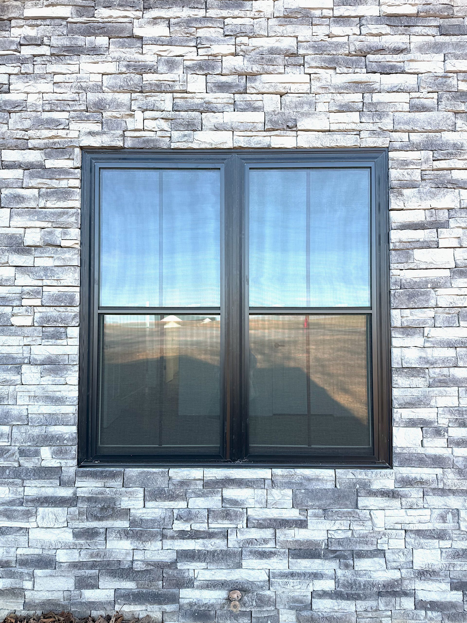 Black-framed window with screen in a gray stone wall. Sunlight reflected in the glass.