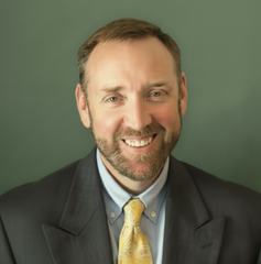 Smiling man in a dark suit and yellow tie against a green background