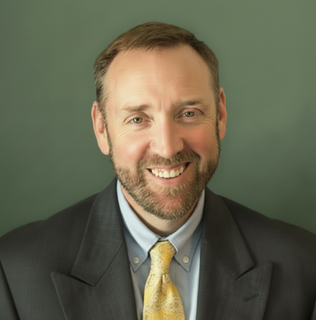 Smiling man in a dark suit and yellow tie against a green background