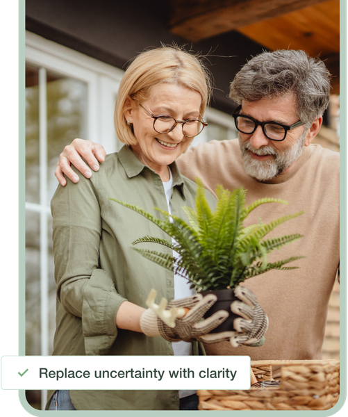 Two people smiling at a potted fern in a bright home, one wearing gloves and holding the plant
