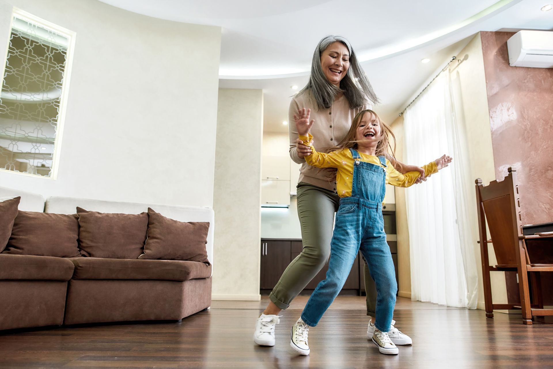 Two people dancing in a bright living room with a brown sofa and wooden floor