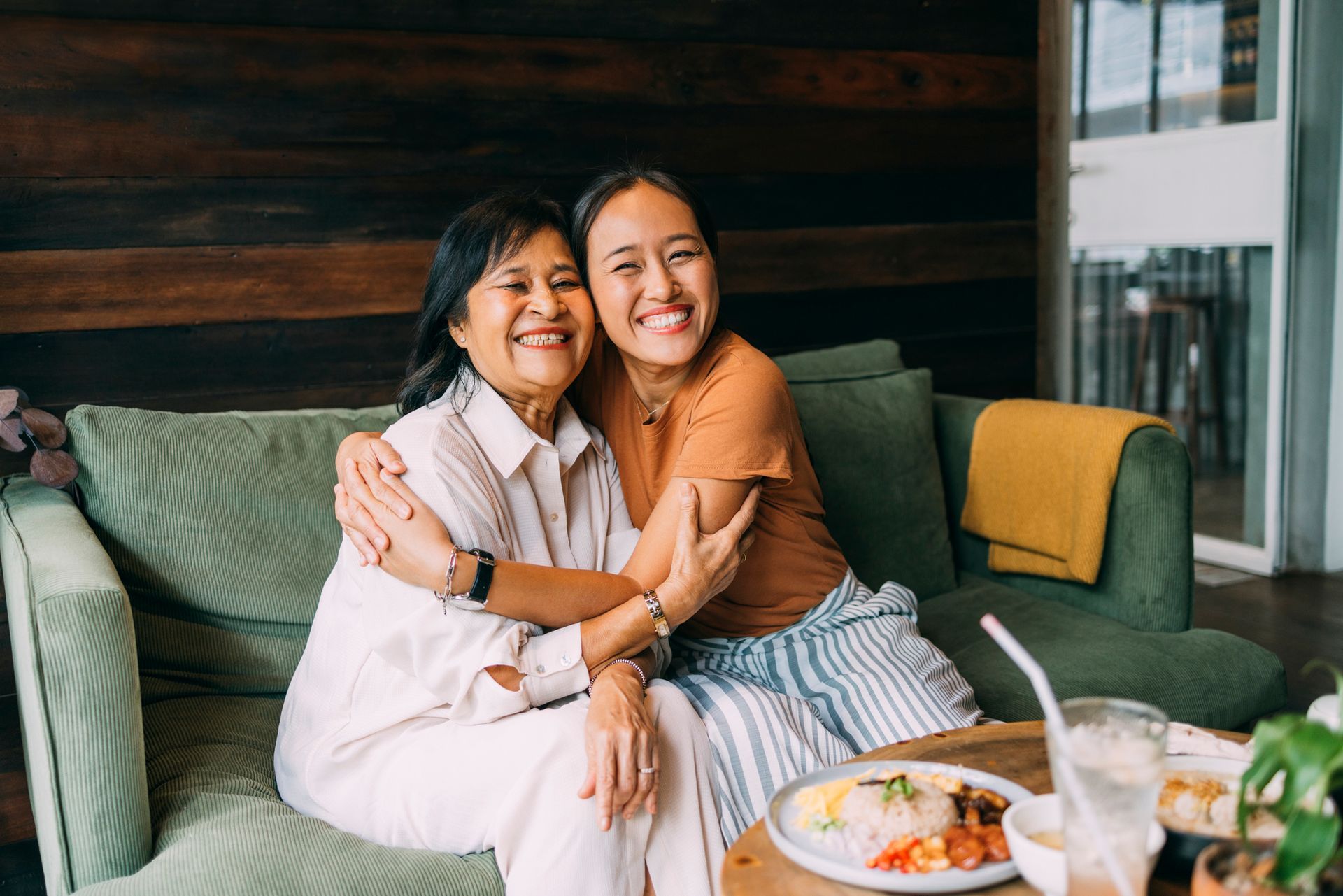 Two people smiling and hugging on a green couch in a cozy café, with plates and drinks on the table