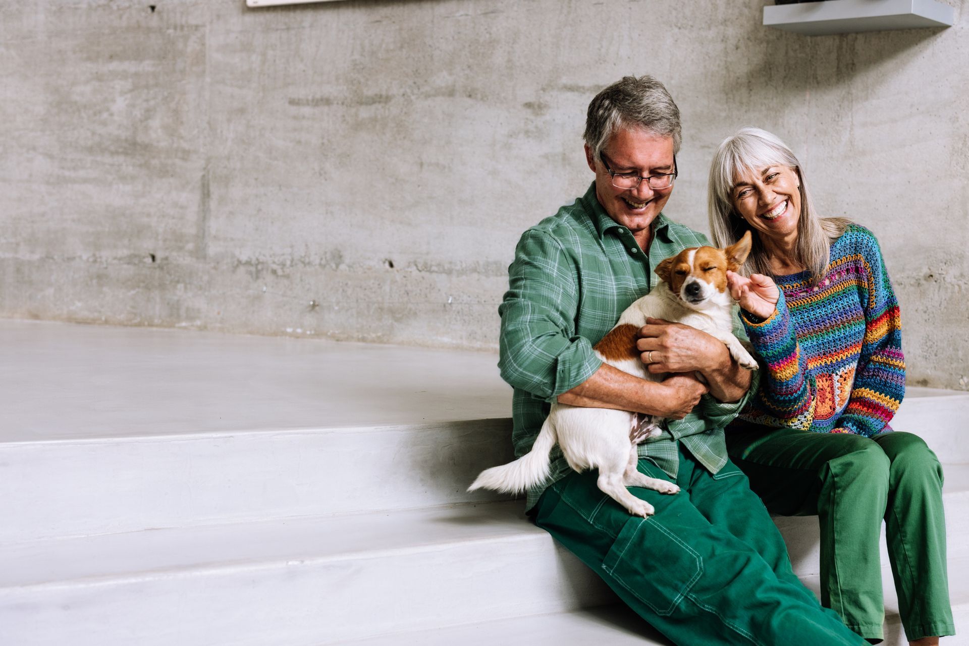 Two people sitting on steps, smiling while holding a small dog, against a concrete wall.