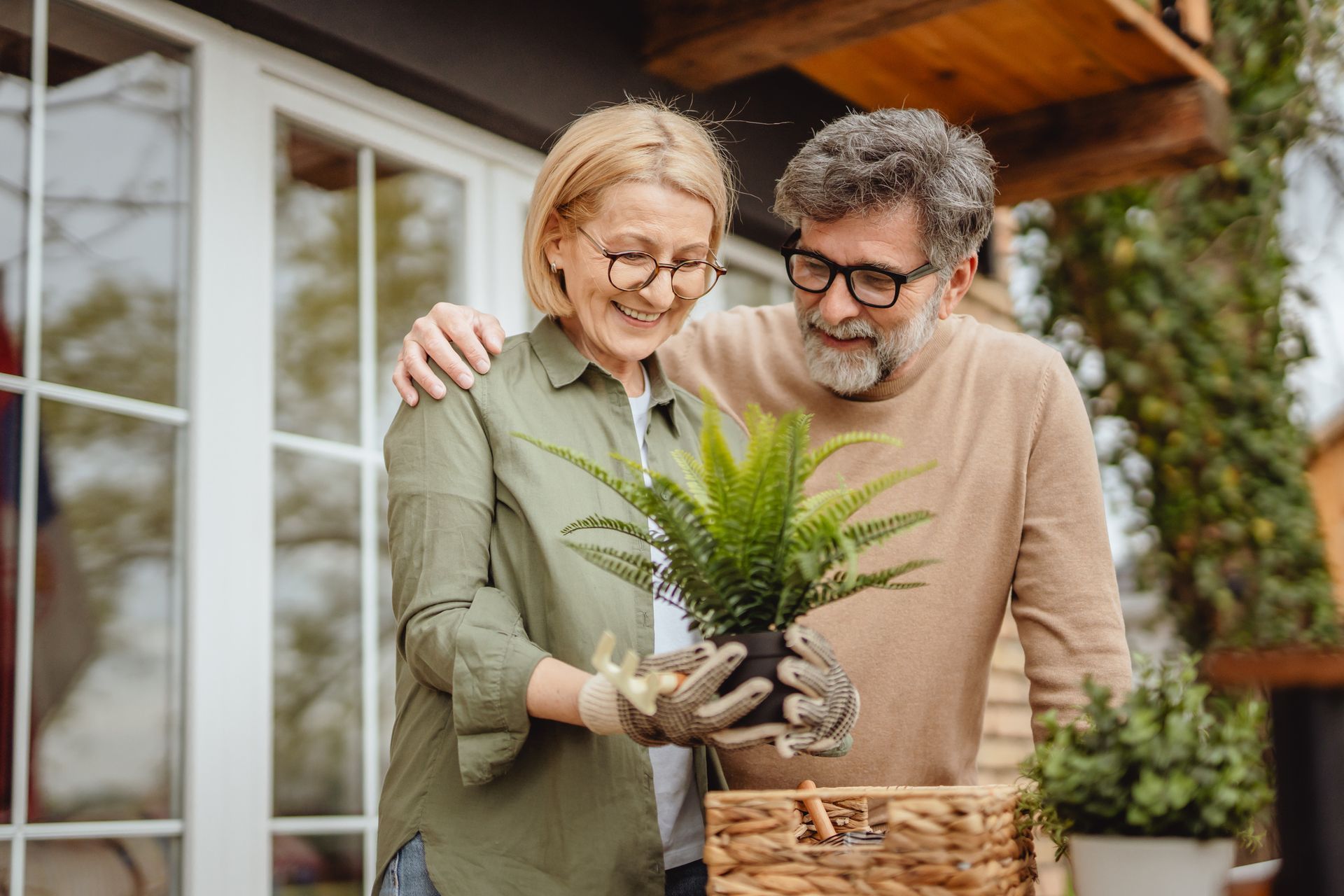 Two people smiling while holding a potted plant beside a wicker basket on a porch