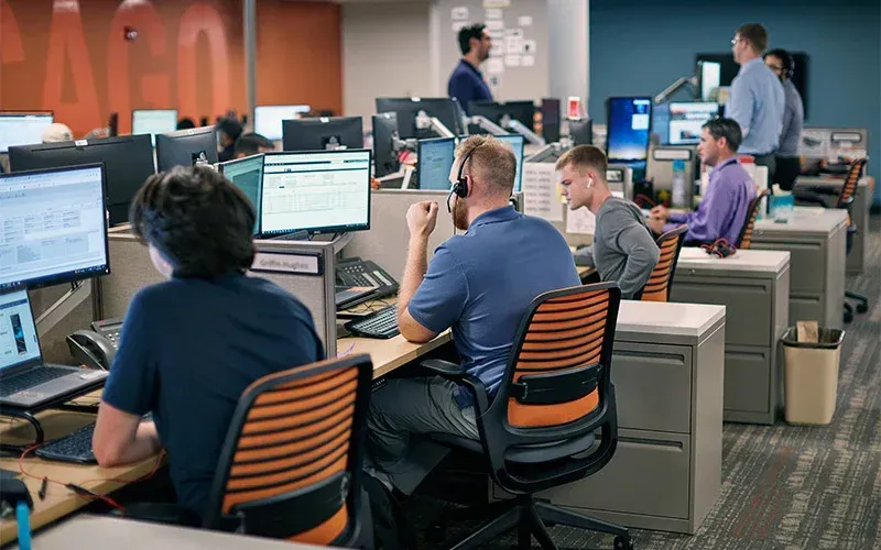 Office with workers at desks, some on headsets, facing computer screens. Bright orange accent wall.